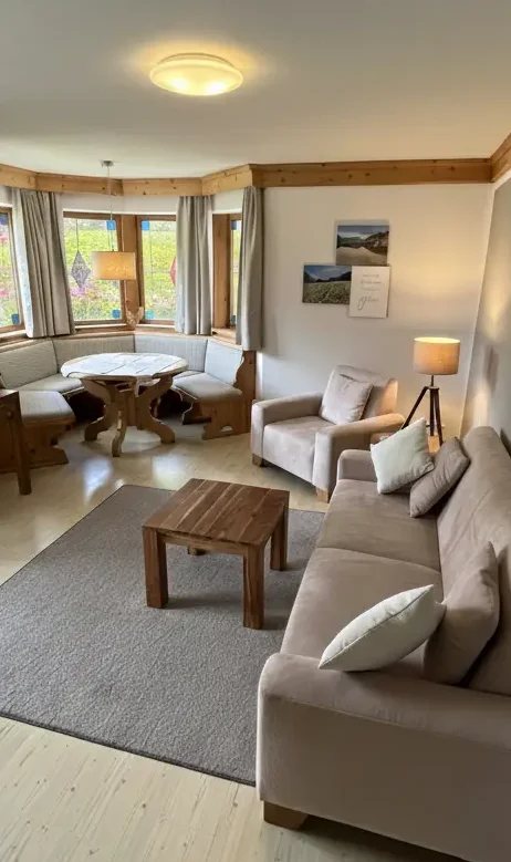 Wohnzimmer-Ferienwohnung-Bergveilchen-Haus-am-Wiesbach-Reit-Im-Winkl Helles und barrierefreies Wohnzimmer mit beigen Sofas, Holztisch und großen Fenstern und Alpenblick
